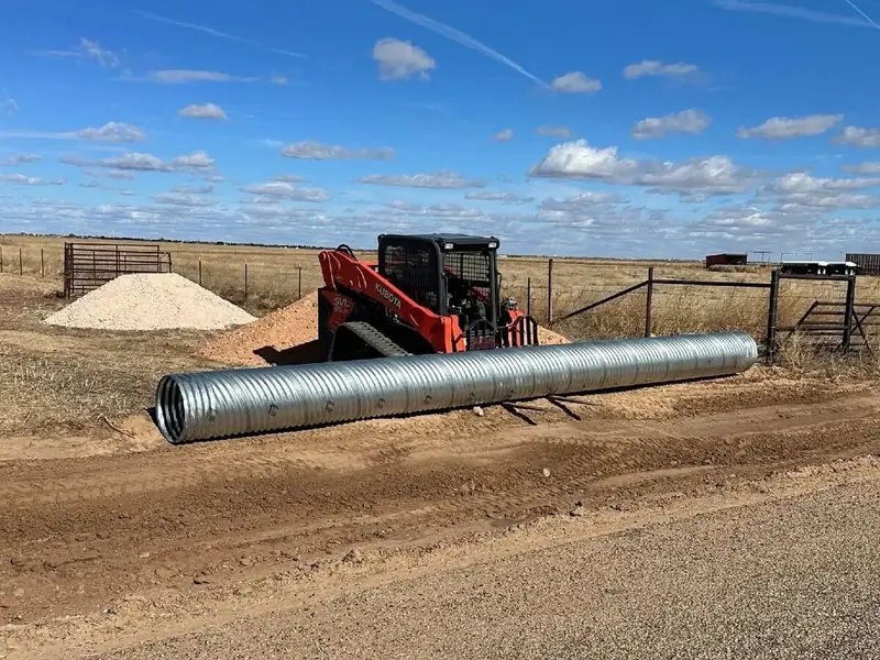 Skid steer carrying a long culvert pipe across a ranch road